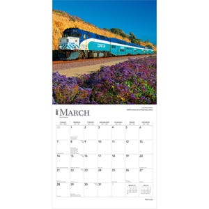 K&ouml;nnte beinhalten: Ein Wandkalender f&uuml;r M&auml;rz 2027 mit einer Fotografie eines blau-wei&szlig;en Coaster-Pendlerzuges, der auf Gleisen neben einem Feld mit violetten Blumen und einem felsigen H&uuml;gel unter einem klaren blauen Himmel entlangf&auml;hrt.