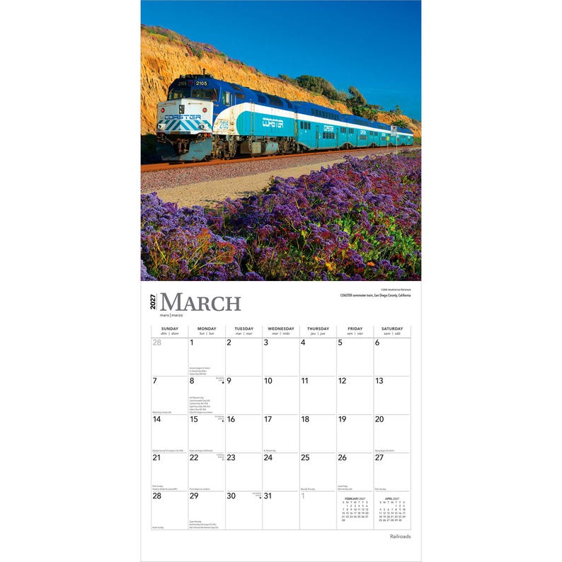 K&ouml;nnte beinhalten: Ein Wandkalender f&uuml;r M&auml;rz 2027 mit einer Fotografie eines blau-wei&szlig;en Coaster-Pendlerzuges, der auf Gleisen neben einem Feld mit violetten Blumen und einem felsigen H&uuml;gel unter einem klaren blauen Himmel entlangf&auml;hrt.