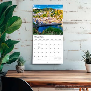 May include: A wall calendar for March 2027 featuring a photograph of a coastal village with white buildings, green hills, and a harbor with boats. The calendar grid displays the days of the month with the text March 2027 clearly visible at the top.