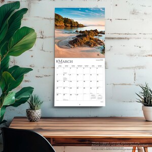 May include: A wall calendar for March 2025 featuring a scenic photograph of a sandy beach with rocky formations, calm water, and a treelined coastline under a blue sky. The calendar grid displays dates, days of the week, and small monthly views.