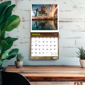 May include: A wall calendar for March 2027 featuring a Banksy graffiti image. The top portion displays a color photograph of a person working on graffiti art near water. The calendar grid is below the image, with the word "MARCH" in green.