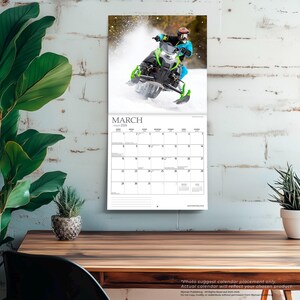 Puede incluir: Calendario de pared para marzo de 2026, con una moto de nieve en acci&oacute;n. La moto de nieve es negra y verde, con una persona que lleva un casco y ropa azul. El calendario es blanco con texto negro.