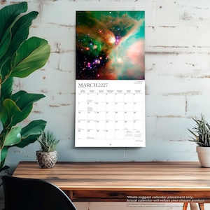 Op de afbeelding: Een wandkalender voor maart 2027 met een levendige afbeelding van een ruimtenevel met wervelende groene, oranje en blauwe kosmische wolken en heldere sterren. Het kalenderraster toont de maand, de dagen van de week en data in zwarte letters.