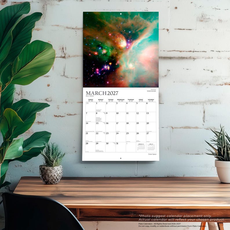 Op de afbeelding: Een wandkalender voor maart 2027 met een levendige afbeelding van een ruimtenevel met wervelende groene, oranje en blauwe kosmische wolken en heldere sterren. Het kalenderraster toont de maand, de dagen van de week en data in zwarte letters.