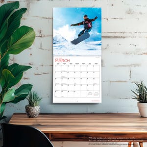 Puede incluir: Un calendario de pared con una fotograf&iacute;a de un snowboarder en el aire sobre un cielo azul brillante y un paisaje de monta&ntilde;a nevado. La p&aacute;gina del calendario muestra el mes de marzo de 2027 con una cuadr&iacute;cula de fechas y el texto MARCH en rojo.