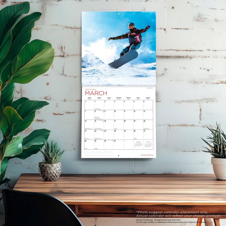 Puede incluir: Un calendario de pared con una fotograf&iacute;a de un snowboarder en el aire sobre un cielo azul brillante y un paisaje de monta&ntilde;a nevado. La p&aacute;gina del calendario muestra el mes de marzo de 2027 con una cuadr&iacute;cula de fechas y el texto MARCH en rojo.