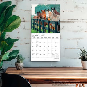 K&ouml;nnte beinhalten: Ein Wandkalender f&uuml;r M&auml;rz 2027 mit einem Foto von vier Pferden hinter einem dunklen Holzzaun auf einer gr&uuml;nen Wiese. Das Kalenderraster zeigt die Tage des Monats, wobei der Text MARCH 2027 deutlich oben auf der Seite aufgedruckt ist.