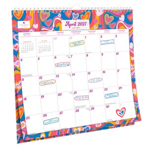Op de afbeelding: Een spiraalgebonden wandkalender voor april 2027, met een kleurrijk psychedelisch ontwerp met harten en wervelingen. De kalender heeft handgeschreven notities voor afspraken en evenementen, waaronder "Meeting" en "Trip to NY."