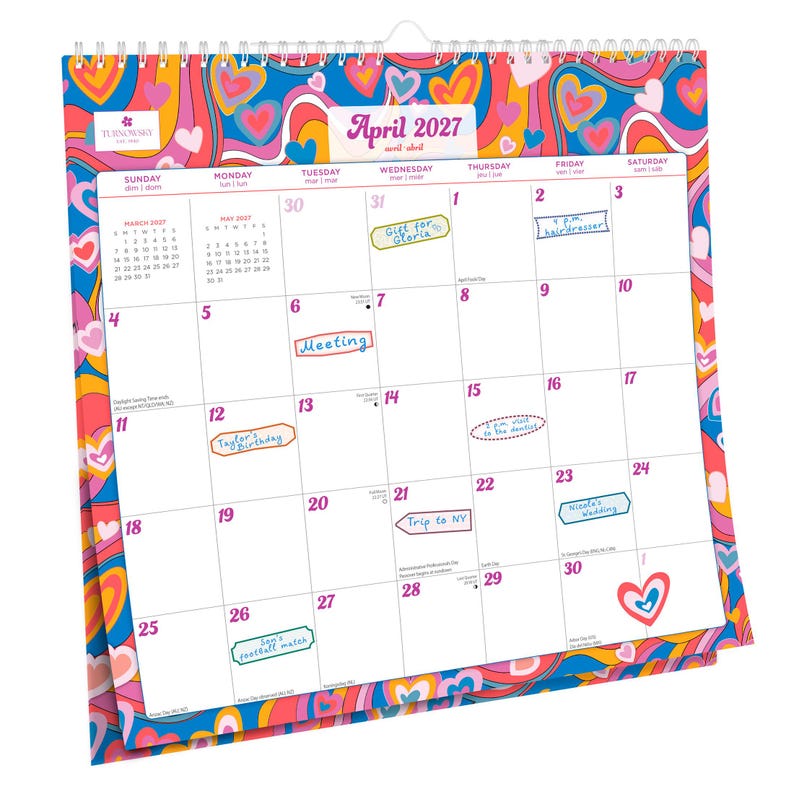 Op de afbeelding: Een spiraalgebonden wandkalender voor april 2027, met een kleurrijk psychedelisch ontwerp met harten en wervelingen. De kalender heeft handgeschreven notities voor afspraken en evenementen, waaronder "Meeting" en "Trip to NY."