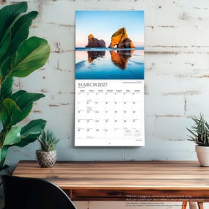 May include: A wall calendar for March 2027 featuring a photograph of large rock formations on a beach at sunset. The calendar grid displays the days of the month with clear black text on a white background, hanging vertically on a white brick wall.