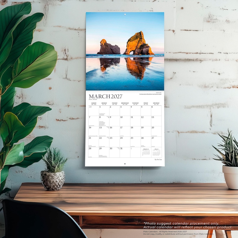 May include: A wall calendar for March 2027 featuring a photograph of large rock formations on a beach at sunset. The calendar grid displays the days of the month with clear black text on a white background, hanging vertically on a white brick wall.