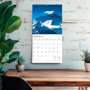 May include: A wall calendar featuring a photograph of a snow-covered mountain under a blue sky. The calendar is open to March, with the month's name at the top and dates below. The calendar is hanging on a white brick wall.