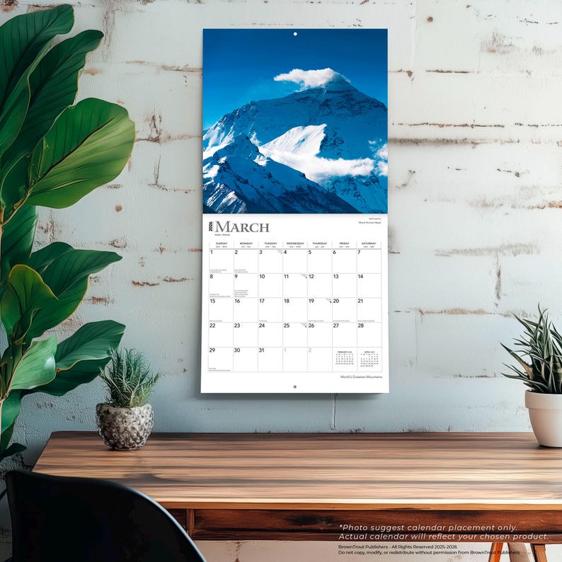 May include: A wall calendar featuring a photograph of a snow-covered mountain under a blue sky. The calendar is open to March, with the month's name at the top and dates below. The calendar is hanging on a white brick wall.