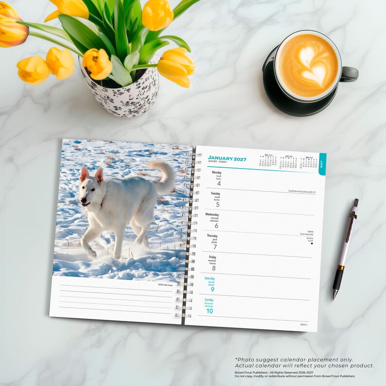 May include: A spiral-bound wall calendar open to January 2027, featuring a photograph of a white dog running in the snow on the left page and a weekly planner layout on the right page, accompanied by a black pen resting on the side.