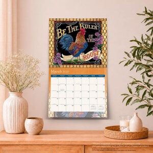 Op de afbeelding: Een wandkalender voor maart 2027 met een kleurrijke haanillustratie en de tekst Be The Ruler Of The Roost. Het kalenderrooster staat onder de afbeelding, die is omlijst door een goudkleurig kippengaaspatroon.