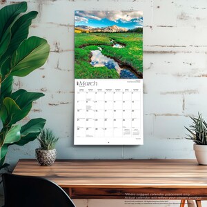 May include: A wall calendar for March 2027 featuring a landscape photograph of a winding stream through a green meadow with mountains in the background. The calendar grid displays the days of the month with the text March 2027 clearly visible at the top.