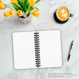 May include: An open spiral bound notebook with lined pages. The notebook is on a white marble surface with a cup of coffee and a pen.