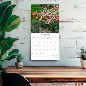 May include: A wall calendar for March 2027 featuring a photograph of cherry blossoms over a pond. The calendar grid displays the month, year, and days of the week. The text "March 2027" is printed at the top of the grid, with "Japanese Gardens" below.