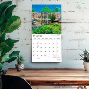 May include: A wall calendar for March 2027 featuring a photograph of a stone waterfall flowing into a pond surrounded by lush green vegetation and a clear blue sky. The calendar grid displays the days of the month with black text on white paper.