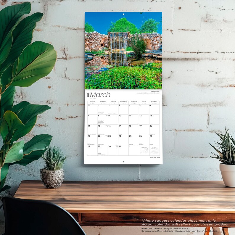 May include: A wall calendar for March 2027 featuring a photograph of a stone waterfall flowing into a pond surrounded by lush green vegetation and a clear blue sky. The calendar grid displays the days of the month with black text on white paper.