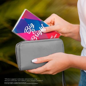 Puede incluir: Una agenda rectangular con un patr&oacute;n geom&eacute;trico vibrante en tonos rosa, azul y morado, con el texto blanco Big & Bright. La agenda se est&aacute; colocando dentro de una cartera gris lisa con cremallera de textura suave y cierre plateado.