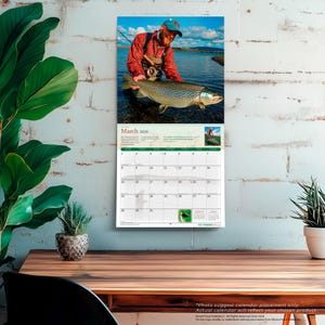 May include: A wall calendar for March 2026 featuring a fisherman holding a large brown trout in a river. The calendar has a grid layout with dates and a small image of a fly fisherman. The calendar is hanging on a white brick wall.