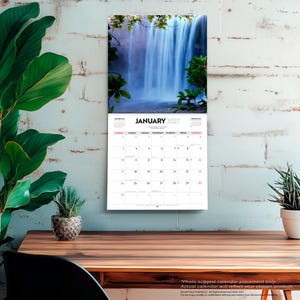 May include: A wall calendar for January 2027 featuring a photograph of a waterfall surrounded by green foliage. The calendar grid displays the month, year, and days of the week with numerical dates in black and red text on a white background.