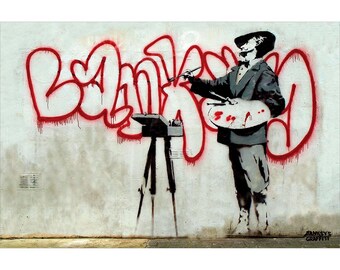 May include: A stencil artwork depicting a figure in a suit and hat, holding a palette and paintbrush, painting the word "BANKSY" in red graffiti on a white wall. A camera is on a tripod. The image is signed "BANKSY'S GRAFFITI".