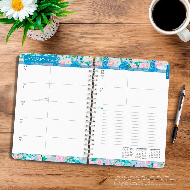 May include: Open spiral-bound planner for 2026, featuring a floral design with blue and pink flowers. The planner is open to a weekly view, with the month of January visible. A pen and a cup of coffee are on the wooden surface.