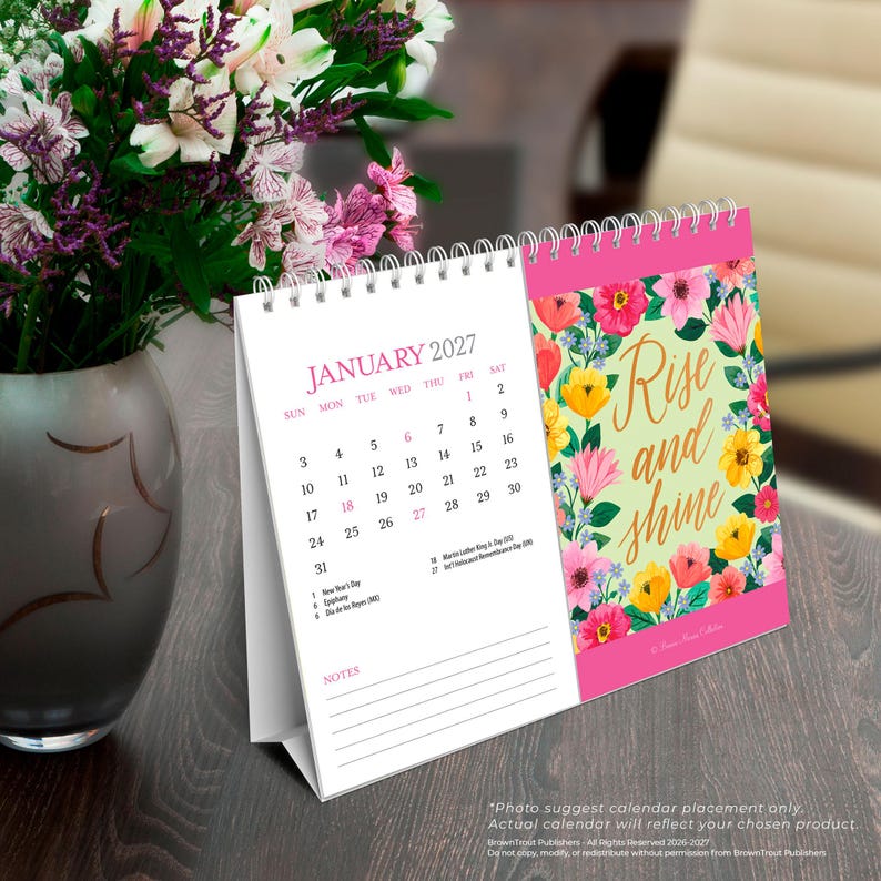 May include: A spiral-bound desk calendar for January 2027 featuring a floral pattern with pink, yellow, and green colors. The right side displays the text Rise and shine in gold script, while the left side shows the monthly grid and notes section.