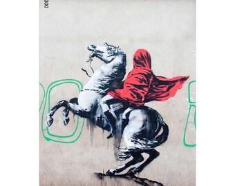 May include: Graffiti art depicting a figure in a red hooded cloak on a white horse. The horse is rearing up. The artwork is on a light beige wall with green graffiti and the text "BANKSY GRAFFITI".