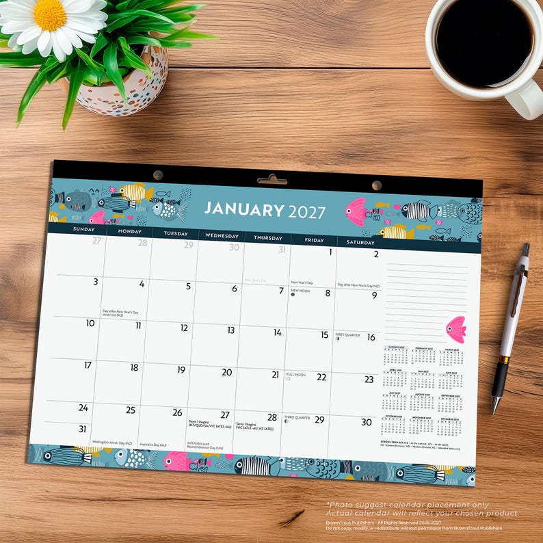 May include: A January 2027 desk calendar featuring a blue border with colorful fish illustrations. The grid displays dates from the 1st to the 31st, with small text for holidays and moon phases. A silver pen rests to the right of the calendar.