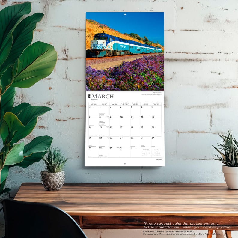 K&ouml;nnte beinhalten: Ein Wandkalender f&uuml;r M&auml;rz 2027 mit einer Fotografie eines blau-wei&szlig;en Coaster-Pendlerzuges, der auf Gleisen neben einem Feld mit violetten Blumen und einem felsigen H&uuml;gel unter einem klaren blauen Himmel f&auml;hrt, mit einem Kalenderraster.