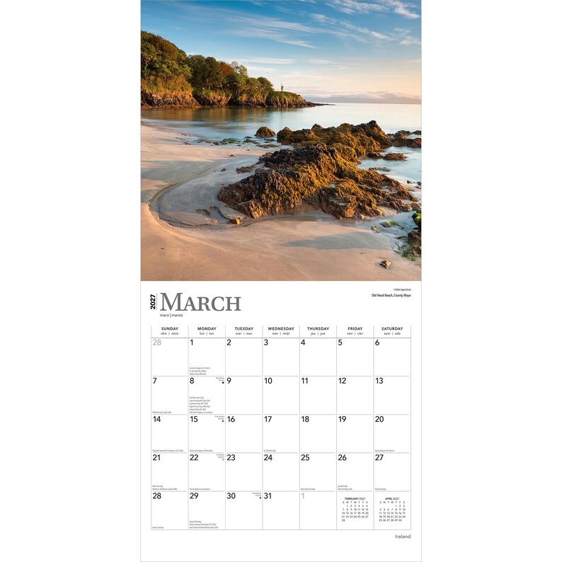 May include: A 2027 wall calendar for the month of March featuring a photograph of a sandy beach with dark, mossy rocks and a tree-lined coastline under a clear sky. The calendar grid displays the days of the month with the text March 2027.