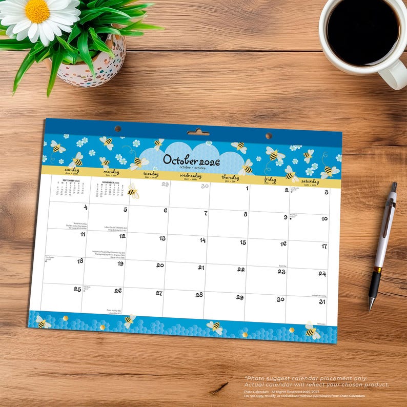 May include: A desk calendar for October 2026 with a blue and yellow bee theme. The calendar has a white background with the days of the month printed in black. A pen, a cup of coffee, and a potted plant are also on the wooden desk.