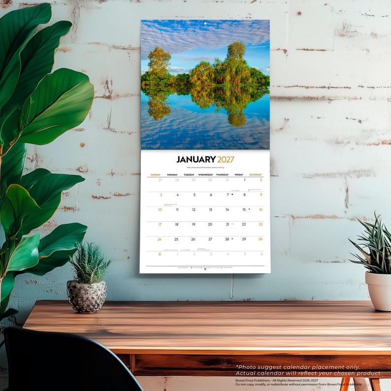 May include: A wall calendar for January 2027 featuring a landscape photograph of trees reflected in calm water. The calendar grid displays the month, year, and days of the week in black text on a white background, with a small hole for hanging.