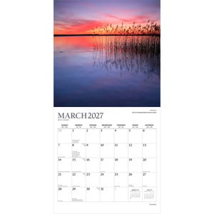 Pu&ograve; includere: Un calendario da parete per marzo 2027 con una fotografia di un tramonto su un lago calmo con alti canneti in primo piano. La griglia del calendario mostra i giorni del mese con un piccolo testo per febbraio e aprile 2027 in basso a destra.