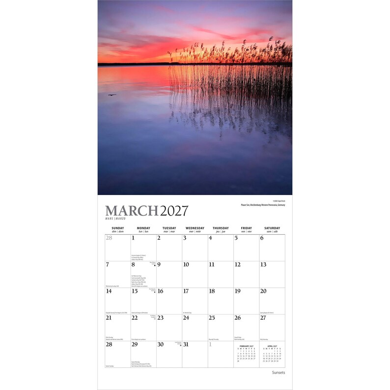 Pu&ograve; includere: Un calendario da parete per marzo 2027 con una fotografia di un tramonto su un lago calmo con alti canneti in primo piano. La griglia del calendario mostra i giorni del mese con un piccolo testo per febbraio e aprile 2027 in basso a destra.
