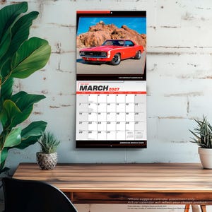 Puede incluir: Un calendario de pared para marzo de 2027 con un Chevrolet Camaro SS rojo brillante de 1969 estacionado frente a un terreno rocoso. La cuadr&iacute;cula del calendario muestra claramente el mes, el a&ntilde;o y las fechas, con el texto American Muscle Cars abajo.