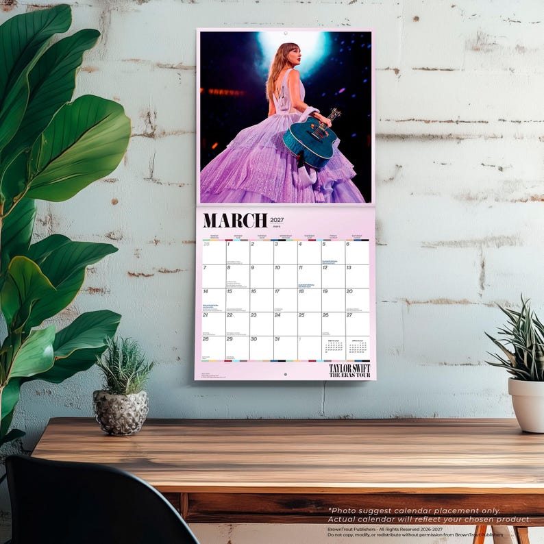 May include: A wall calendar for March 2027 featuring a photograph of a performer in a long, layered lavender gown holding a teal acoustic guitar. The calendar grid displays the month, year, and the text Taylor Swift The Eras Tour at the bottom.
