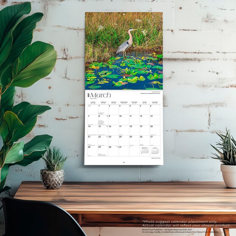 Puede incluir: Calendario de pared con una foto de una garza en un estanque con nen&uacute;fares. El calendario muestra el mes de marzo. El calendario est&aacute; colgado en una pared de ladrillo blanco sobre una mesa de madera.