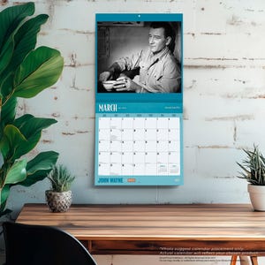 May include: A wall calendar featuring a black and white image of John Wayne. The calendar has a teal border and the month of March is displayed. The words "JOHN WAYNE" are printed at the bottom.