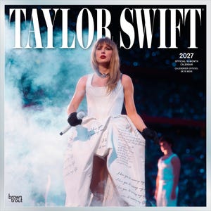 May include: A 2027 official 16-month calendar featuring Taylor Swift on the cover. The image shows the artist in a white dress with handwritten text, black gloves, and a microphone, surrounded by blue stage smoke and a blurred figure.