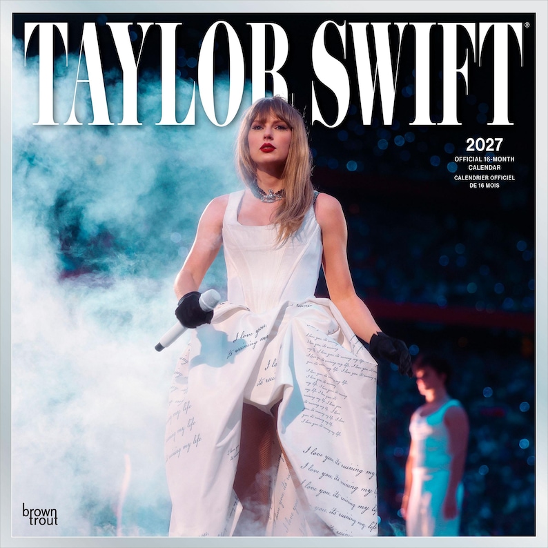 May include: A 2027 official 16-month calendar featuring Taylor Swift on the cover. The image shows the artist in a white dress with handwritten text, black gloves, and a microphone, surrounded by blue stage smoke and a blurred figure.