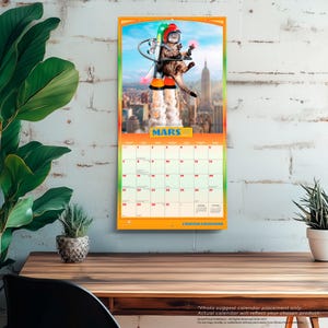 May include: A wall calendar for the month of March featuring a cat wearing a red helmet and a jetpack flying over a city skyline. The calendar grid displays the days of the month with the text MARS and CHATONS GROGNONS printed on the page.