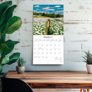May include: A wall calendar for March 2027 featuring a photograph of a dirt path winding through a field of white flowers under a blue sky with clouds. The calendar grid displays the days of the month with the month name and year at the top.