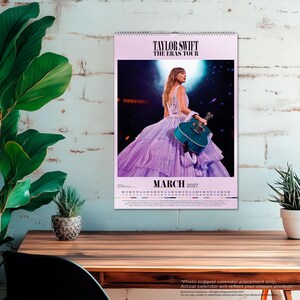 Puede incluir: Un calendario de pared para marzo de 2027 con Taylor Swift The Eras Tour. La imagen muestra a una persona en el escenario con un vestido lavanda brillante de varias capas, sosteniendo una guitarra ac&uacute;stica azul verdoso bajo un foco. La cuadr&iacute;cula est&aacute; debajo.