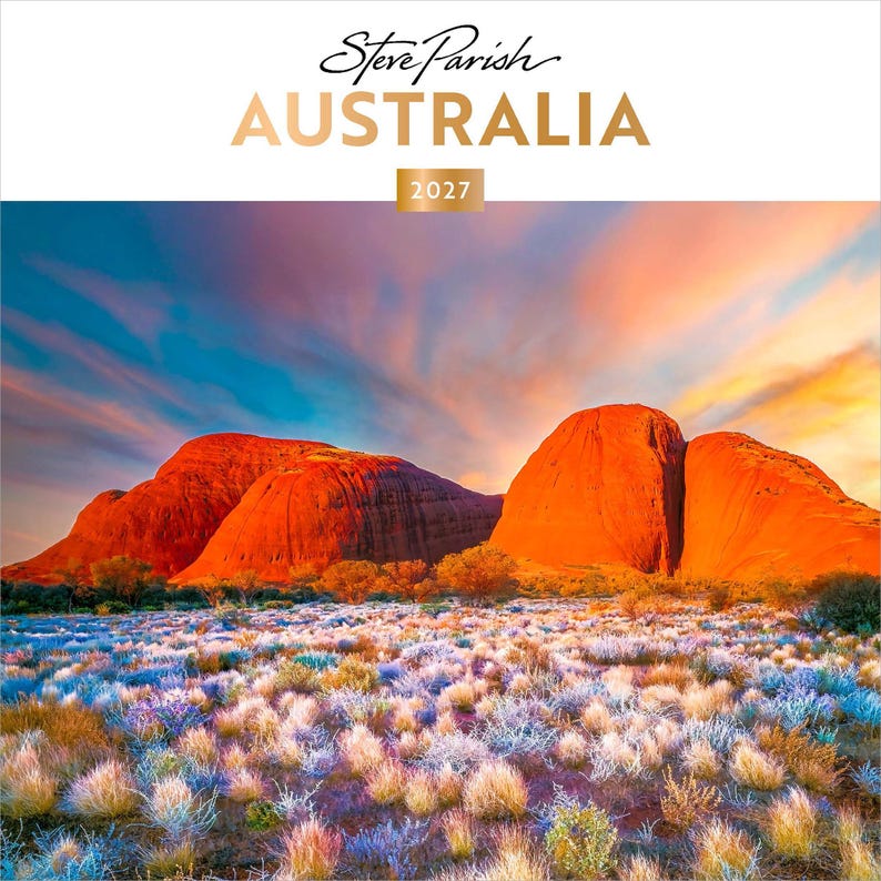 May include: A 2027 wall calendar titled Steve Parish AUSTRALIA featuring a landscape photograph of large red rock formations under a sunset sky with orange, pink, and blue clouds, above a field of low-lying desert shrubs and grasses.