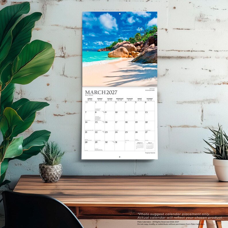 May include: A wall calendar for March 2027 featuring a photograph of a tropical beach with turquoise water, white sand, and large brown rock formations under a blue sky with white clouds. The calendar grid displays dates in black text on white paper.