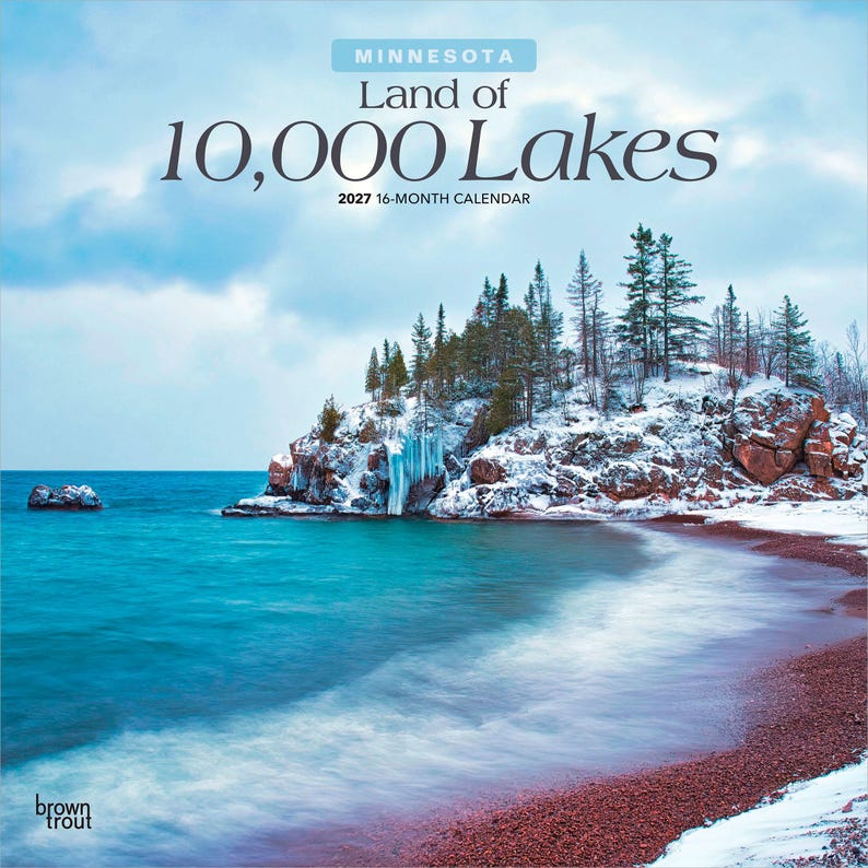 May include: A 2027 16-month calendar titled Minnesota Land of 10,000 Lakes. The cover features a winter landscape with a rocky, snow-covered shoreline, evergreen trees, and icy blue water meeting a reddish-brown pebble beach under a cloudy sky.